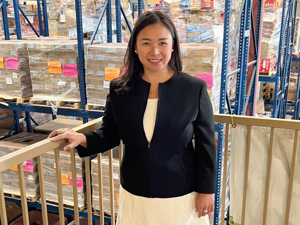 Image: Debbie Espinosa stands in a warehouse in front of food-filled storage racks.