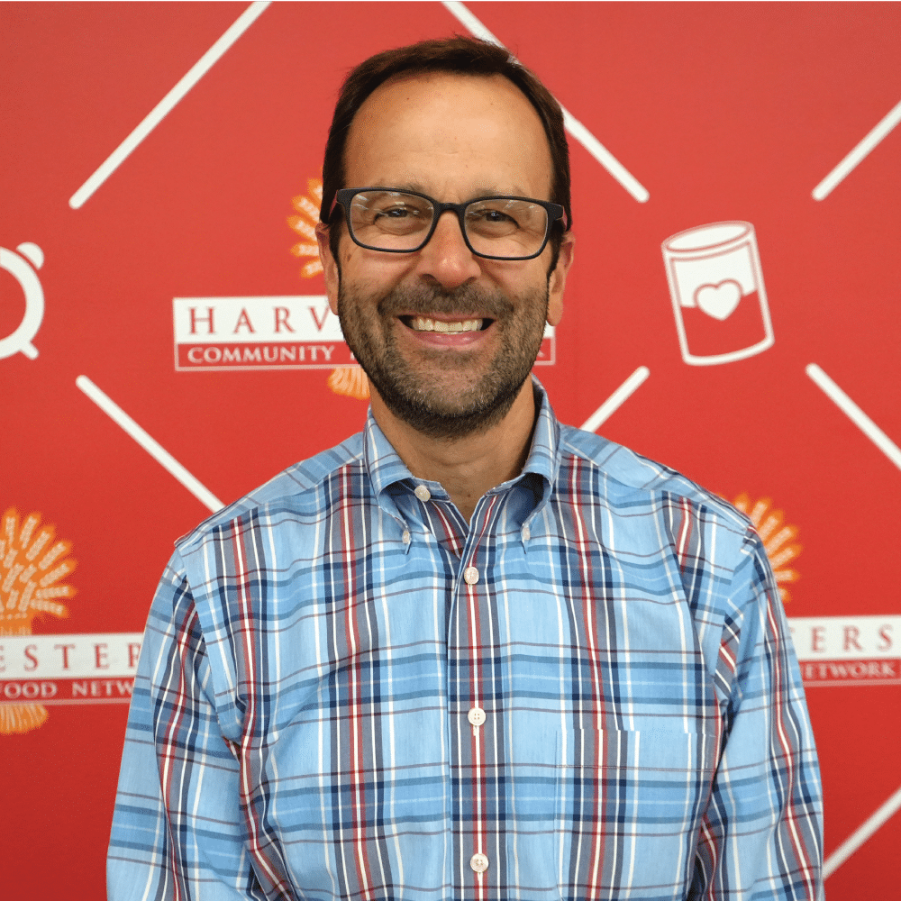 Image: Chirs Krantz, Harvesters board member, smiles in front of a Harvesters-branded backdrop. He is wearing a blue plaid shirt and glasses.
