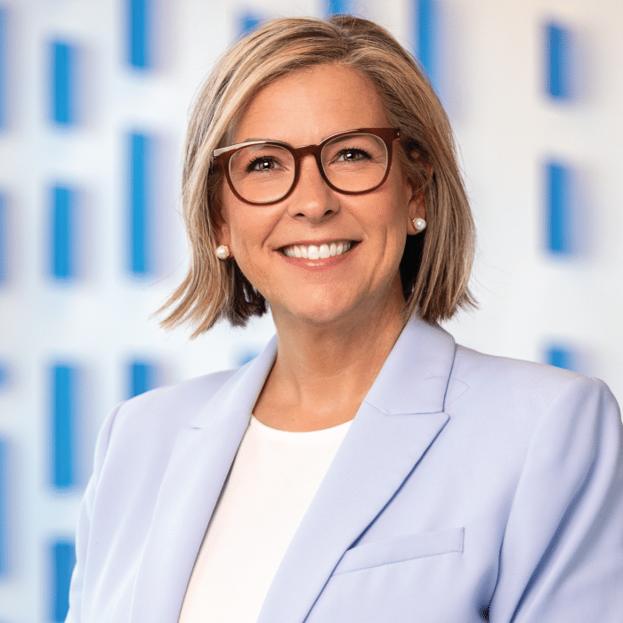 Harvesters Board Member Christa Dubil wears a light blue blazer in front of a blue and white background.