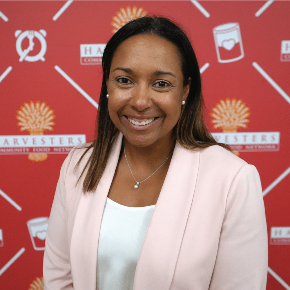 Image: Kamera Meaney, Harvesters board member, smiles in front of a Harvesters-branded backdrop. She is wearing a pink blazer over a white shirt and a gold necklace.