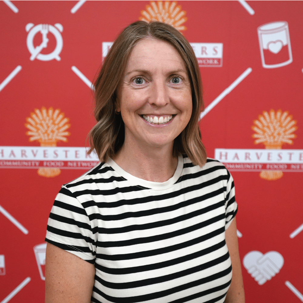 Image: Lisa Herren, Harvesters board member, smiles in front of a Harvesters-branded backdrop. She is wearing a black and white striped shirt.