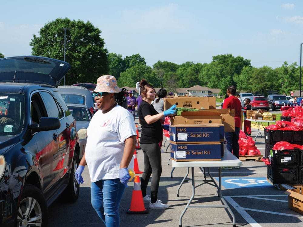 Volunteers load boxes of fresh food into waiting vehicles during a drive-through food distribution. A volunteer wearing gloves walks alongside a vehicle while others stack produce and meal kits on tables in a parking lot on a sunny day.