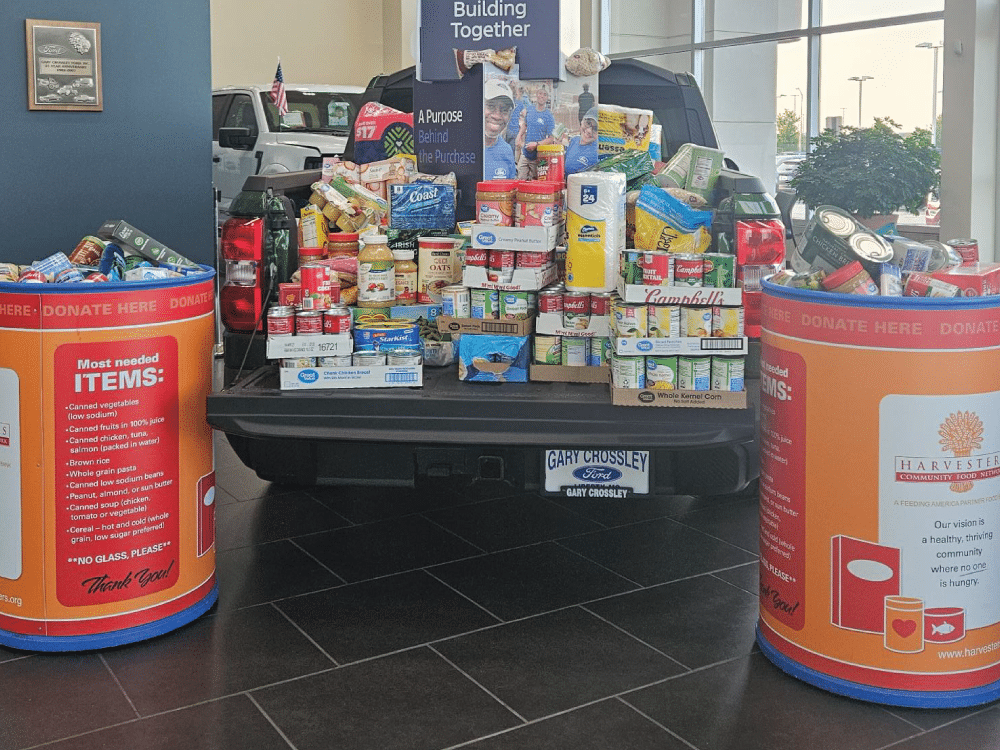 Food donations fill a pickup truck bed between two Harvesters donation barrels.