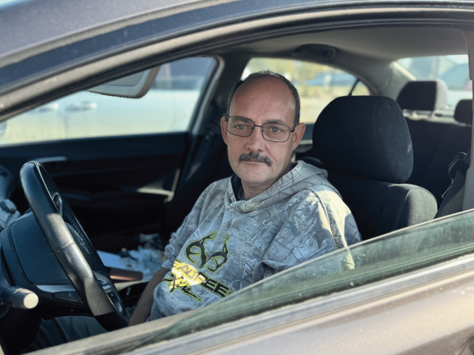 Bill sits in the driver's seat of his car with the window down, looking toward the camera. He wears glasses and a camouflage hoodie, with sunlight illuminating his face as the car's interior and a blurred parking lot are visible in the background.