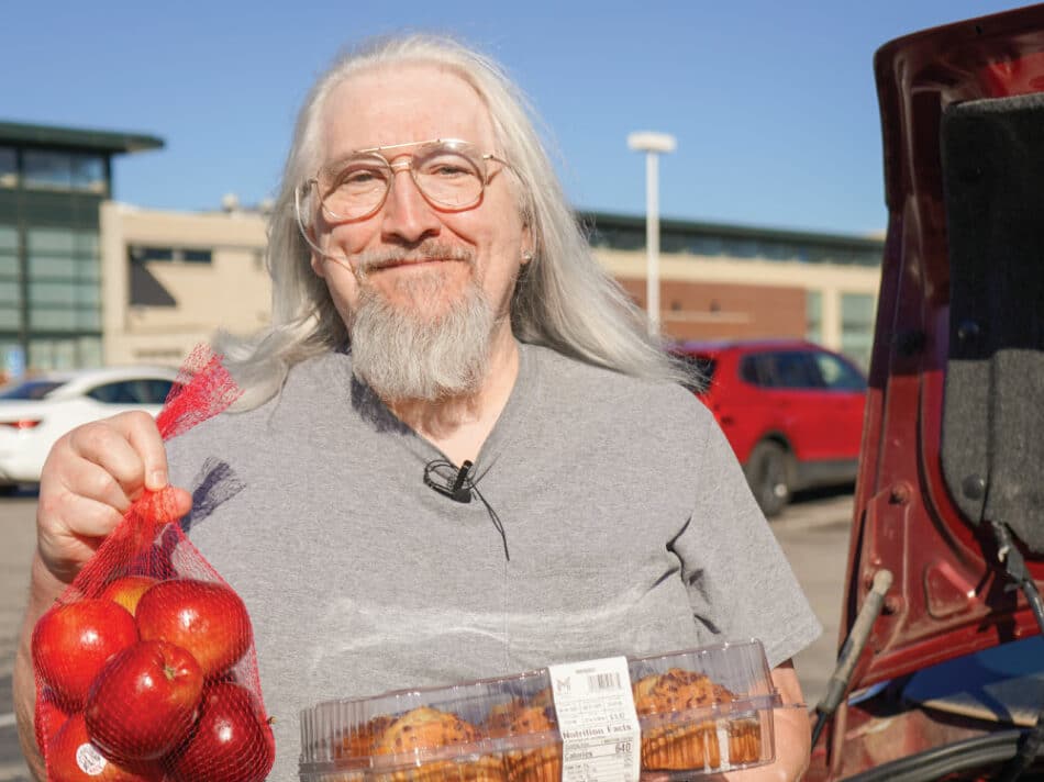 A smiling person with long gray hair and glasses stands beside an open vehicle, holding a bag of red apples and a container of muffins while receiving food assistance in a parking lot on a sunny day.