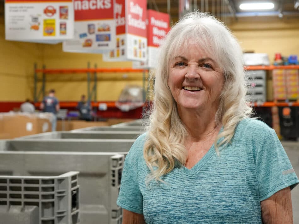 Harvesters Donor Ann Marie Stands in a blue shirt in the sorting area of the website.