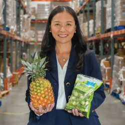 Harvesters President and CEO, Debbi Espinosa, stands smiling in an aisle of the warehouse in a blue blazer holding a pineapple in one hand and a bag of Brussel sprouts in the other.