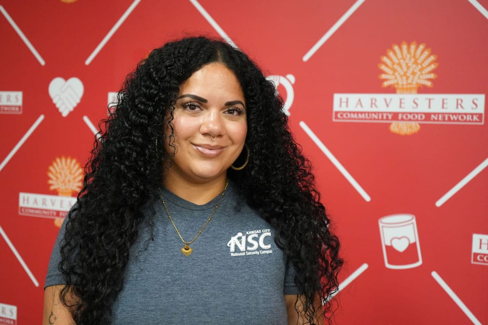 Woman wearing a Kansas City National Security Campus shirt smiles in front of a red Harvesters backdrop.