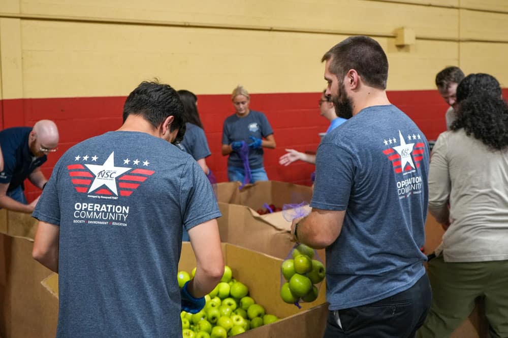 Volunteers in Operation Community shirts pack green apples into purple mesh bags at Harvesters.