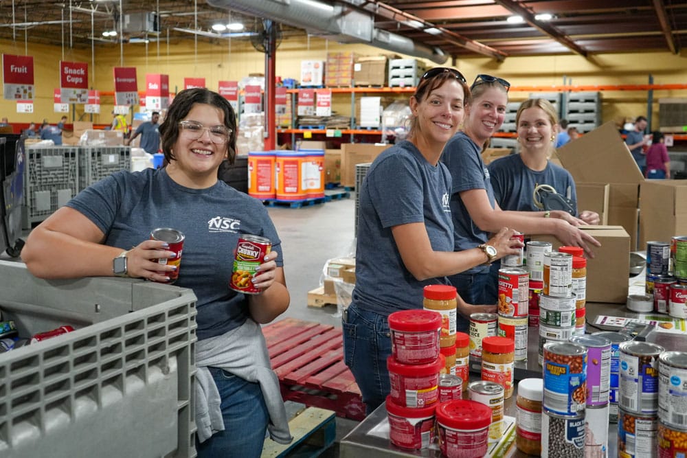 Four smiling volunteers sort canned goods and peanut butter jars at a table in the Harvesters warehouse.
