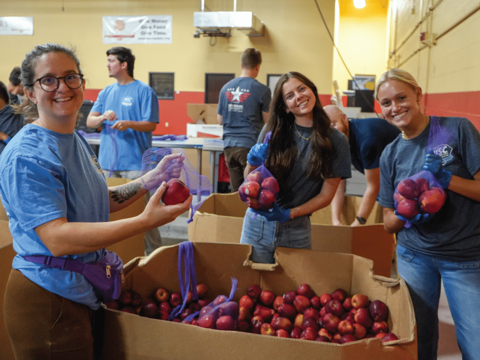 Three smiling volunteers sort red apples into purple mesh bags beside a large bin of apples in a Harvesters warehouse.